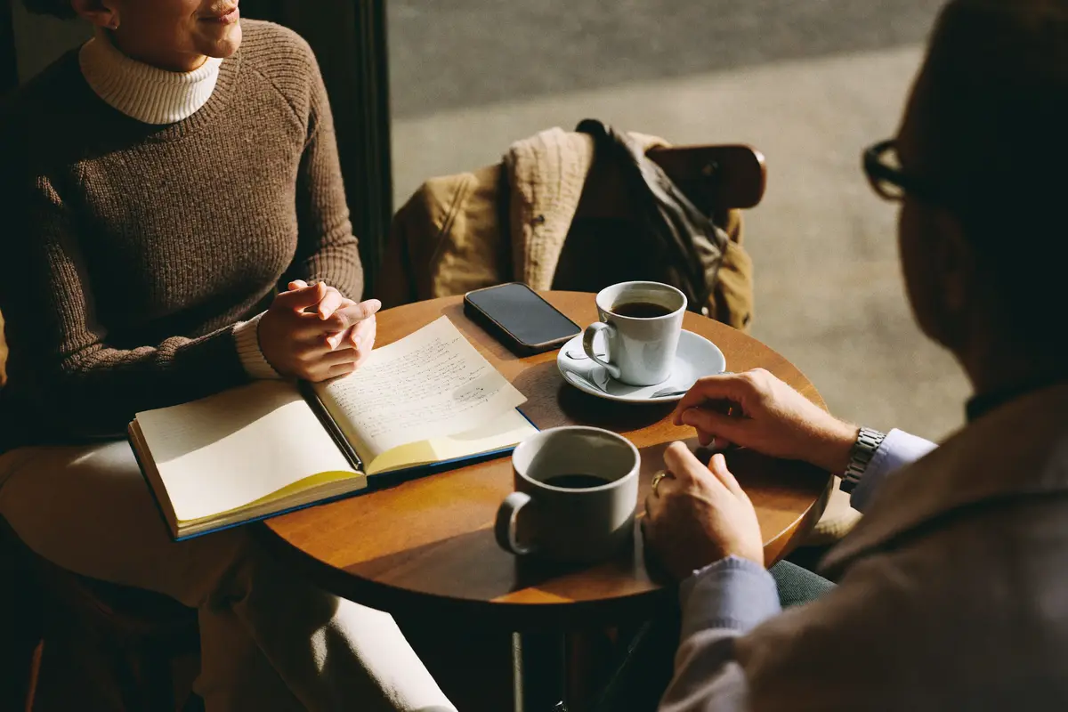 Two people talking over coffee at a small cafe table with a notebook