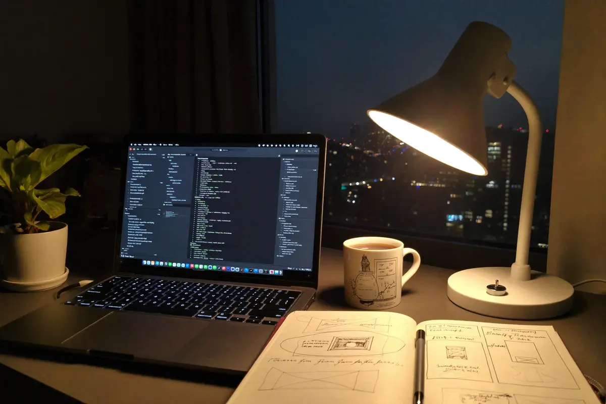 A desk at night with a laptop showing a code editor, a lamp, coffee, and a notebook of website sketches
