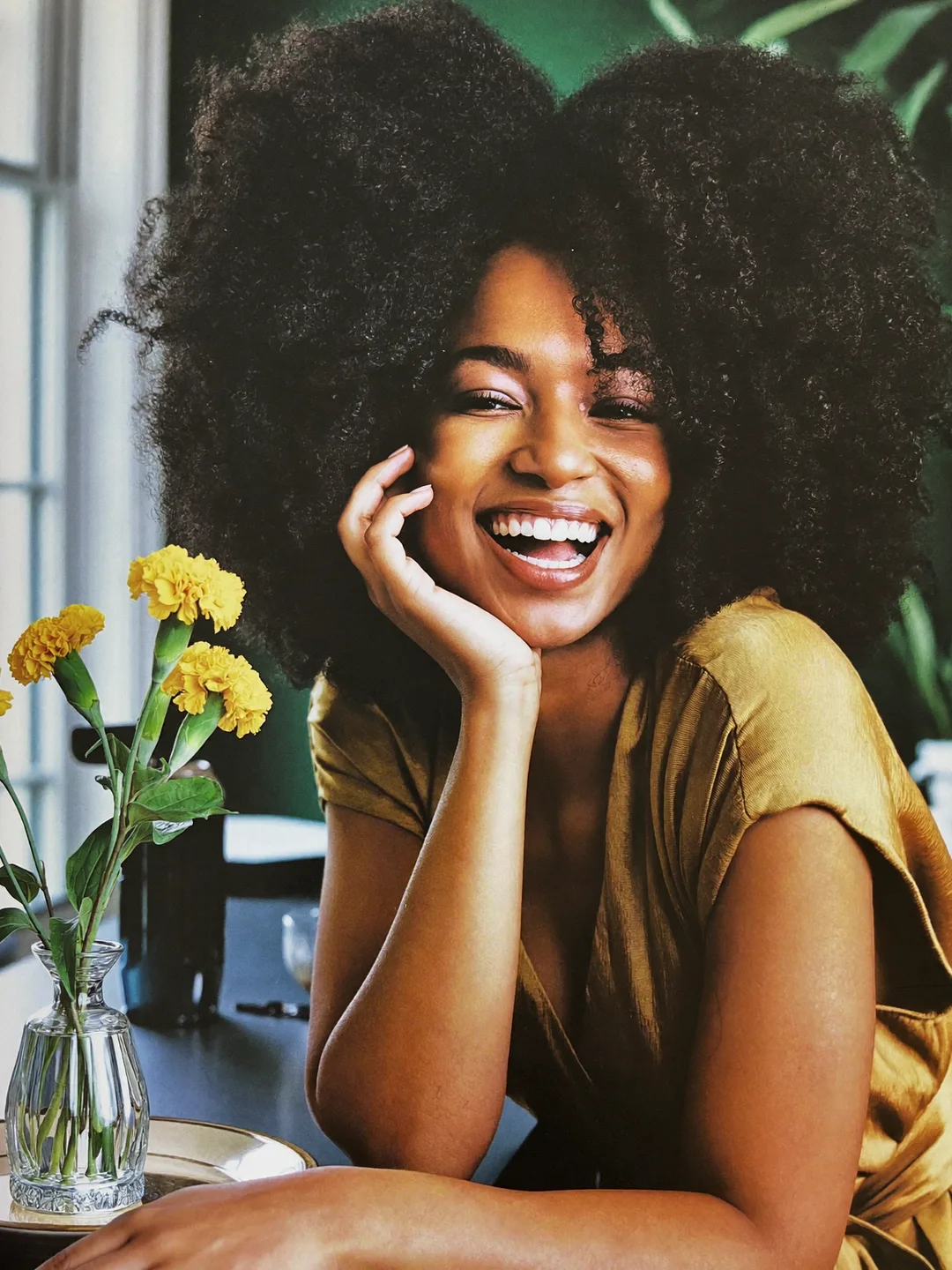 Portrait of stylist Amara laughing, natural curls, mustard silk top, marigolds on the console beside her