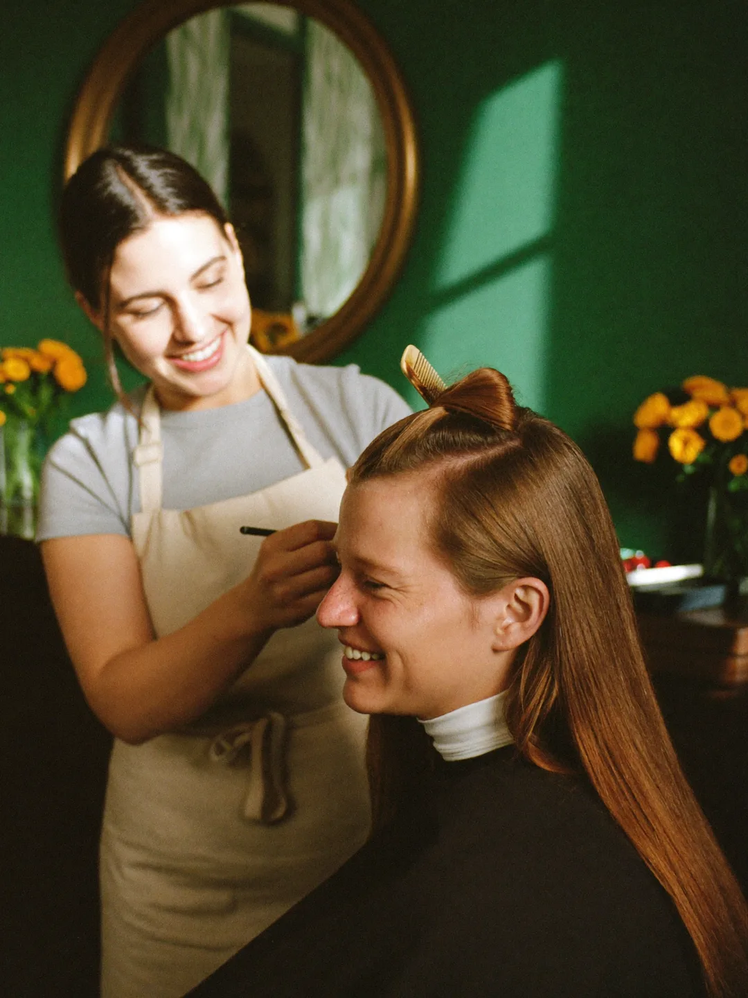Stylist sectioning a client's hair in warm window light, both smiling mid-conversation