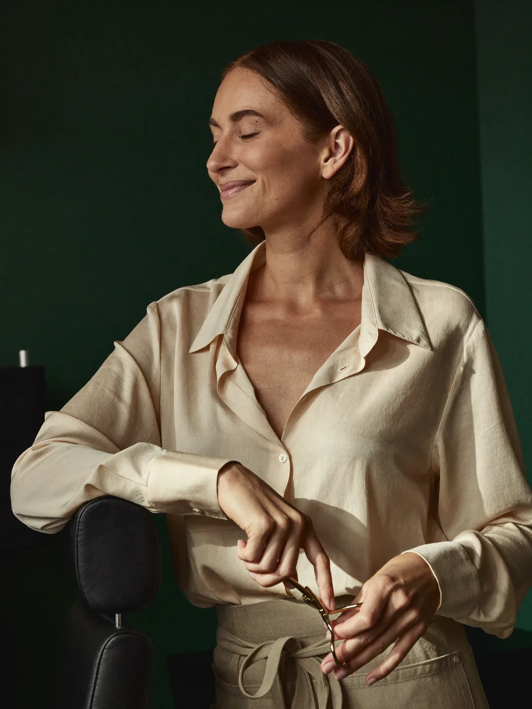 Portrait of salon owner Wren, auburn bob, cream blouse and linen apron, beside her styling chair